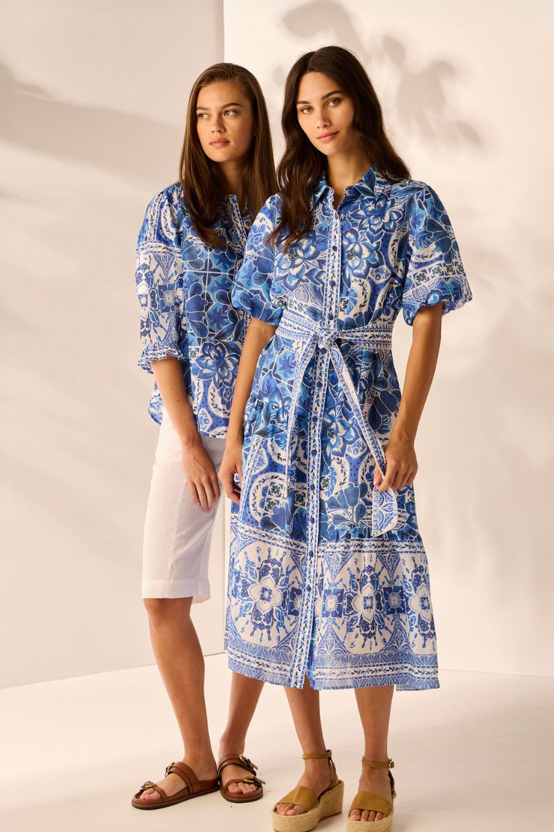 Two women one wearing blue patterned dress the other blue patterned shirt standing against a white background