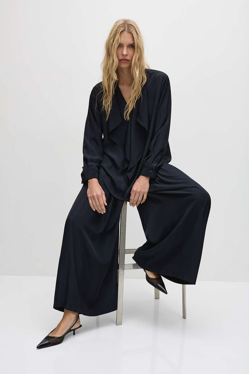 Woman wearing Mela Purdie Maxi Ribbon Shirt F67 8696 and Soft Vent Pant F67 1676 in black sitting on a stool against a white background