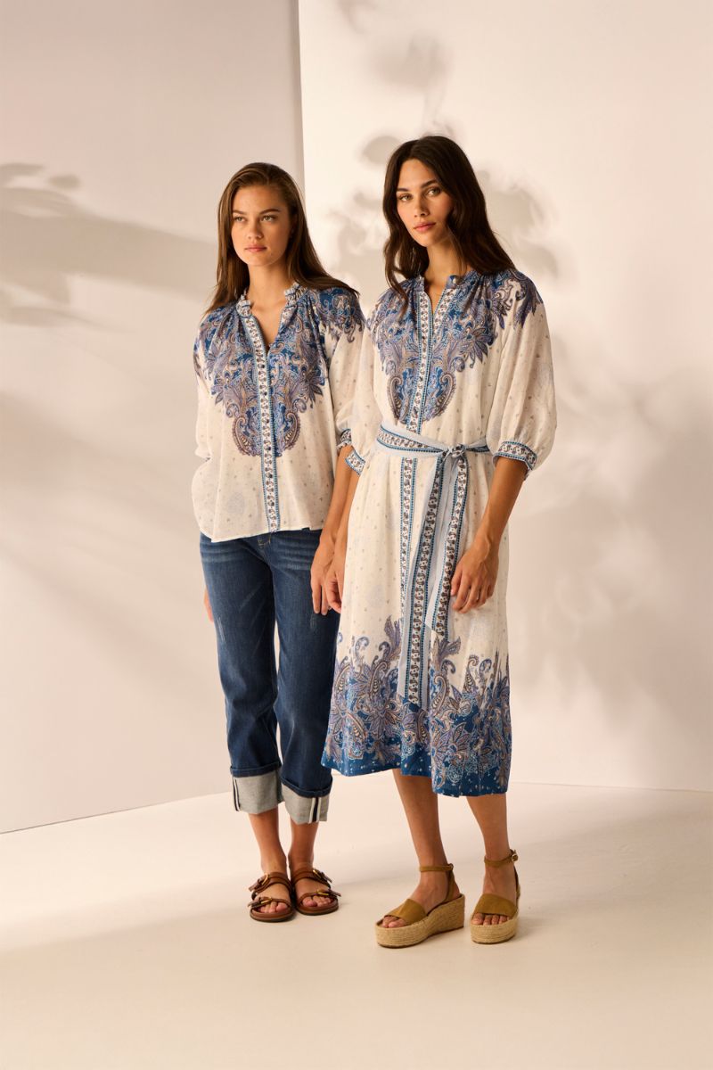 Two women wearing white and blue patterned outfits against a plain background