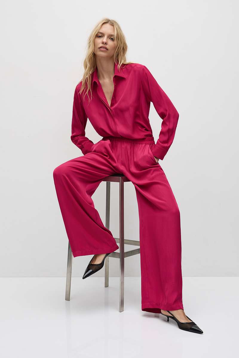 Woman wearing Mela Purdie Soft Shirt F67 2822 and Soft Vent Pant F67 1676 in Fig sitting on a stool against a white background
