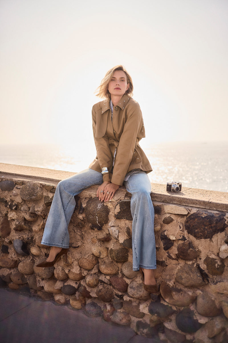 Person sitting on a stone wall wearing a Cotton Twill Short Trench with Belt and Rhinestones Detail jacket and Light Blue Goldie Joseph Ribkoff jeans.