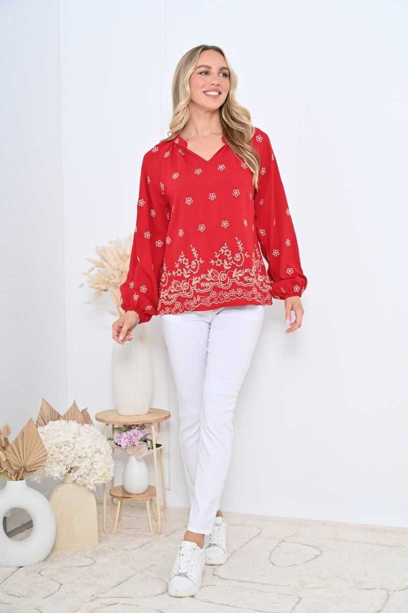 Woman wearing a red embroidered blouse and white pants in a minimalistic room.