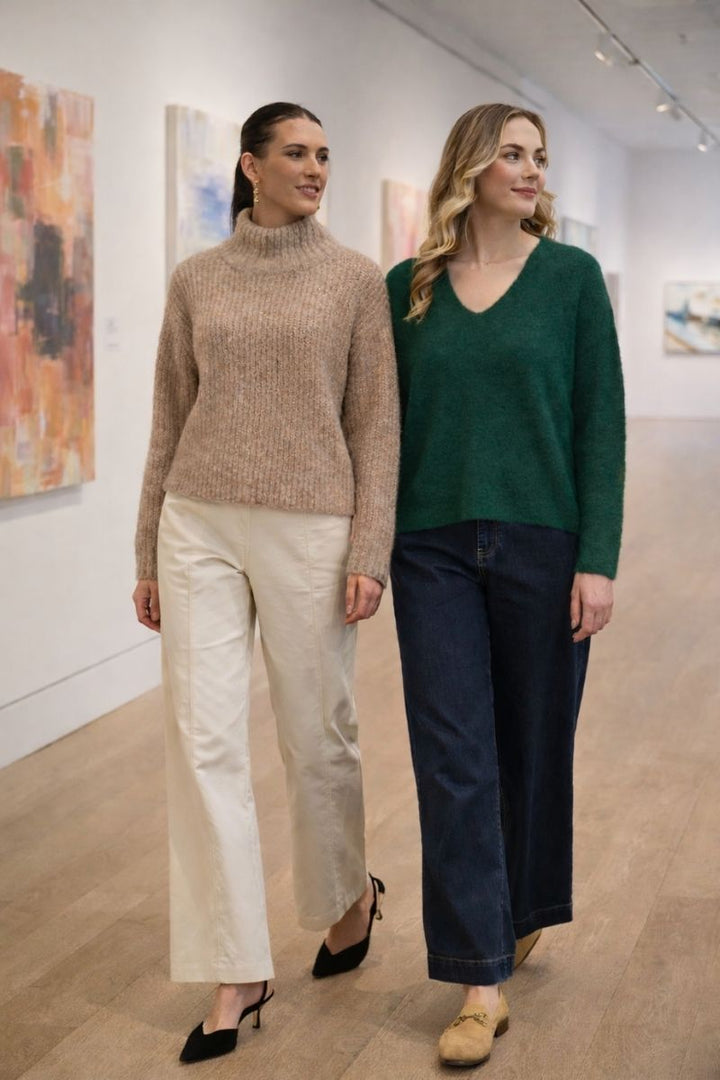 Two women wearing foil sweaters in green with a v neck  and cream with a high roll neck walking in an art gallery
