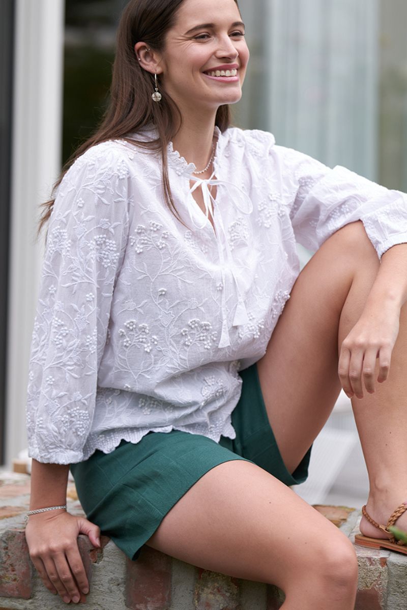 Woman wearing a white lace top and green shorts sitting outdoors.