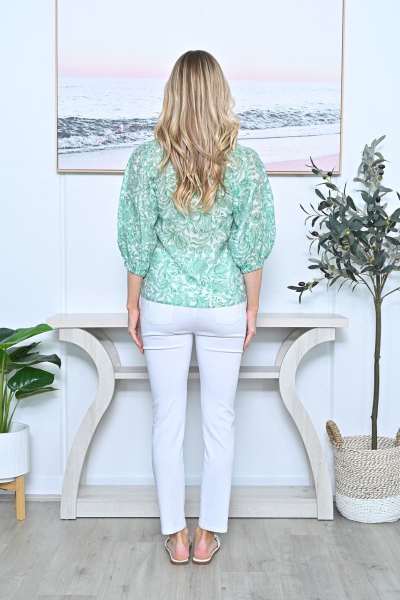 Back view of Woman wearing a green patterned top and white pants standing in a room with a decorative console table and plants.