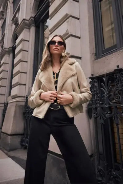 Woman wearing a Joseph Ribkoff beige  coat with a faux fur trim and Black Goldie wide leg jeans in sunglasses standing in front of an urban building.