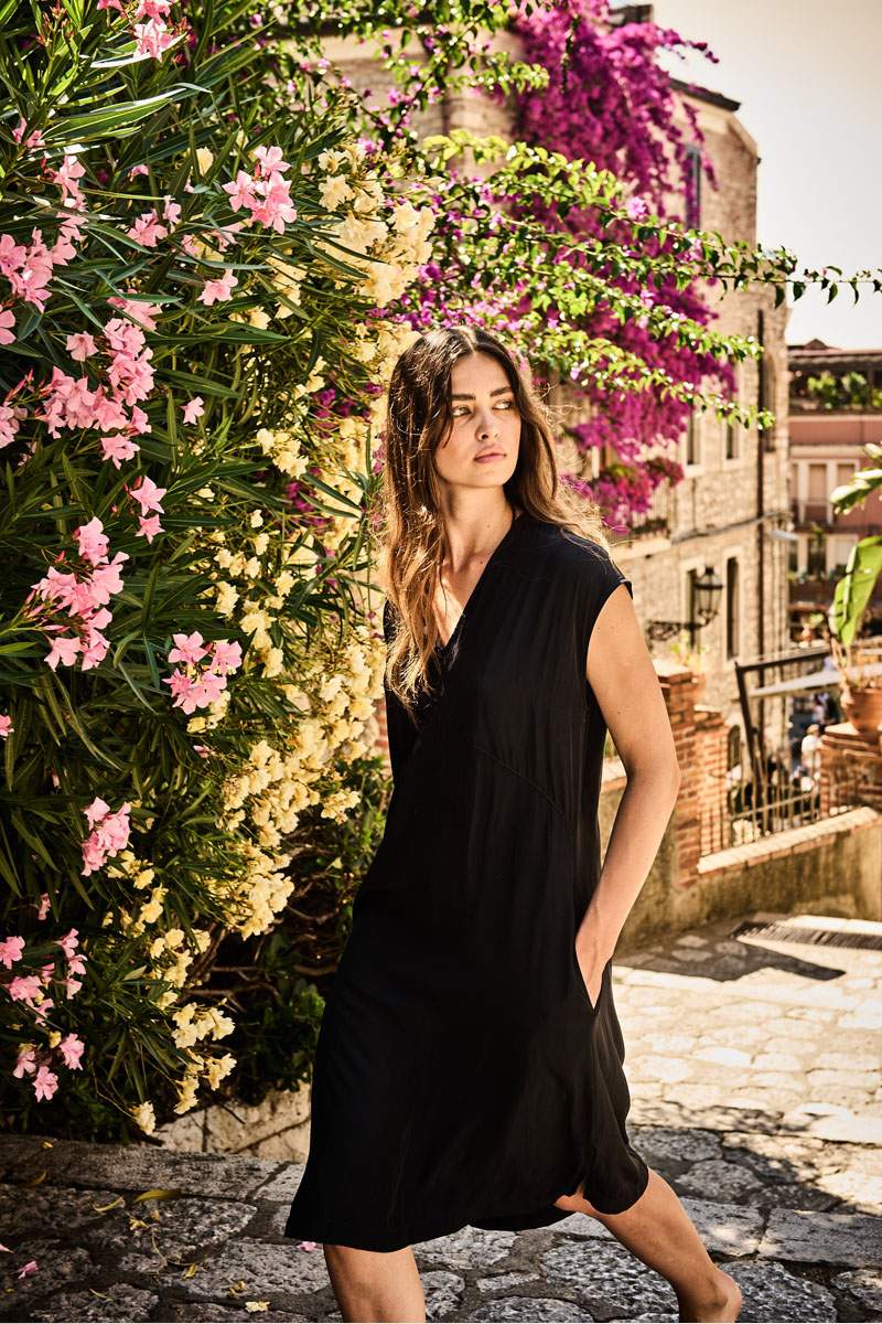 Woman wearing Black Mela Purdie Mache dress walking in a european street with Flowers and cobblestone streets on a sunshine day. Shop mela purdie at weekends.
