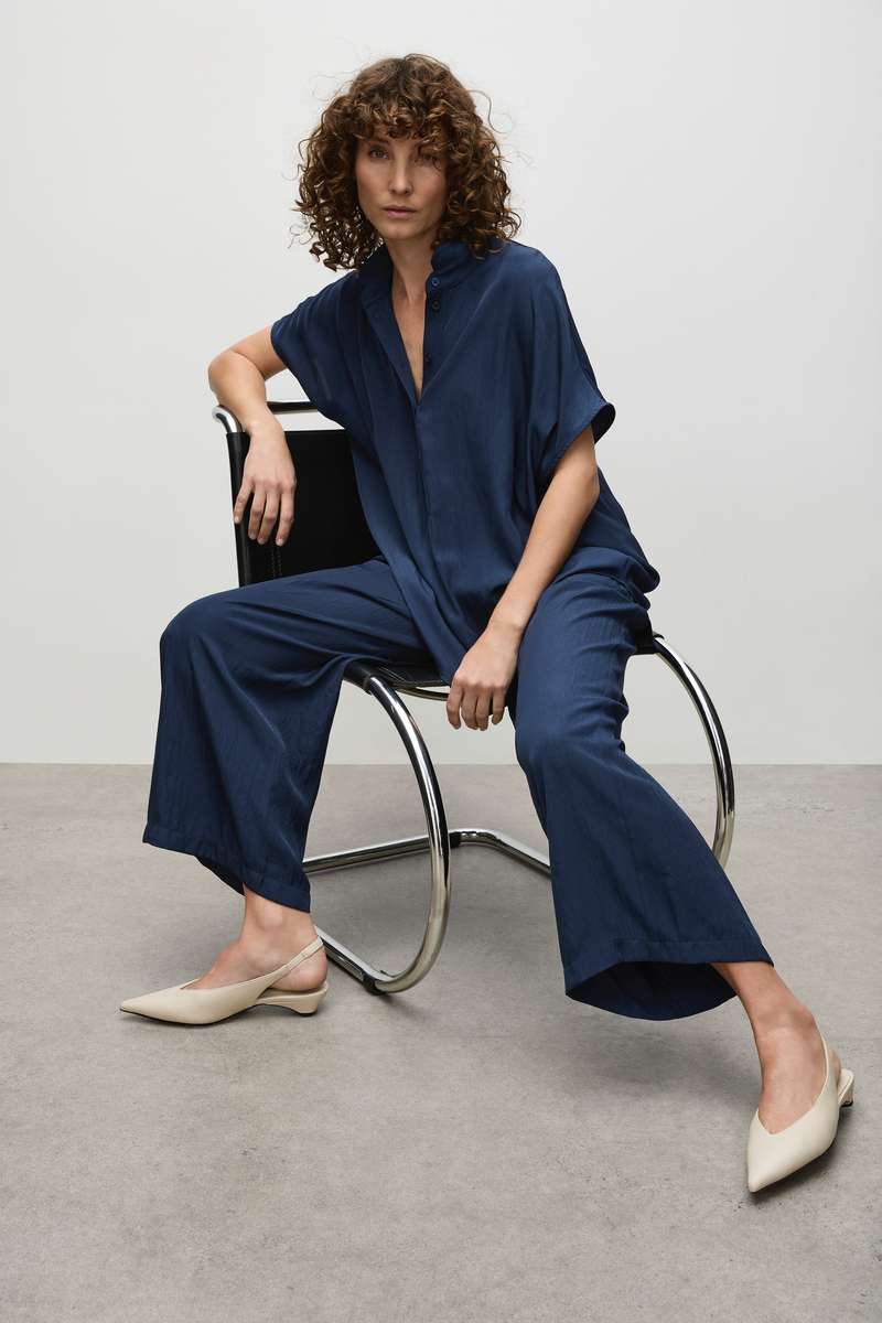 Model Wearing Mela Purdie Tab Stand Shell F67 8646 and Pace Pant F67 1740  in Orient seated on chair on cement floor against white background