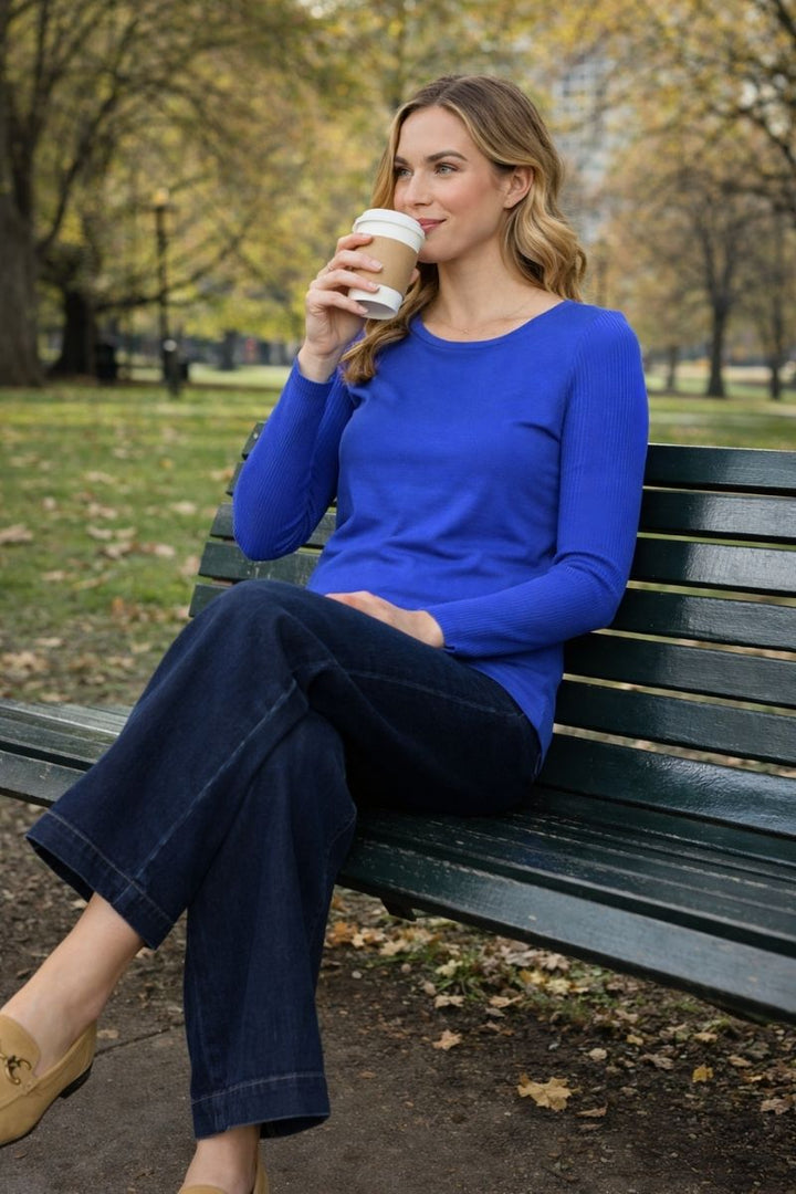 Woman wearing electric blue sweater and dark blue jeans by new zealand label foil clothing sitting on a bench in a park drinking coffee