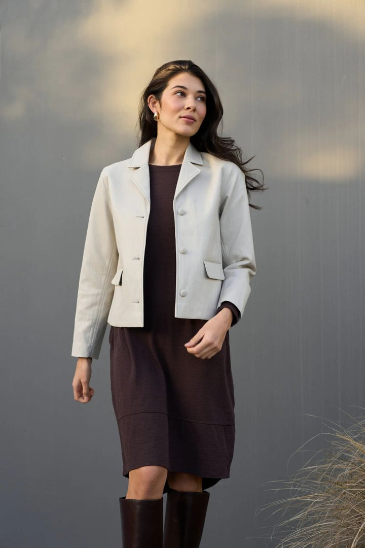 Woman wearing the faux real jacket by foil clothing in Stone over a dark dress with brown boots against a gray wall.