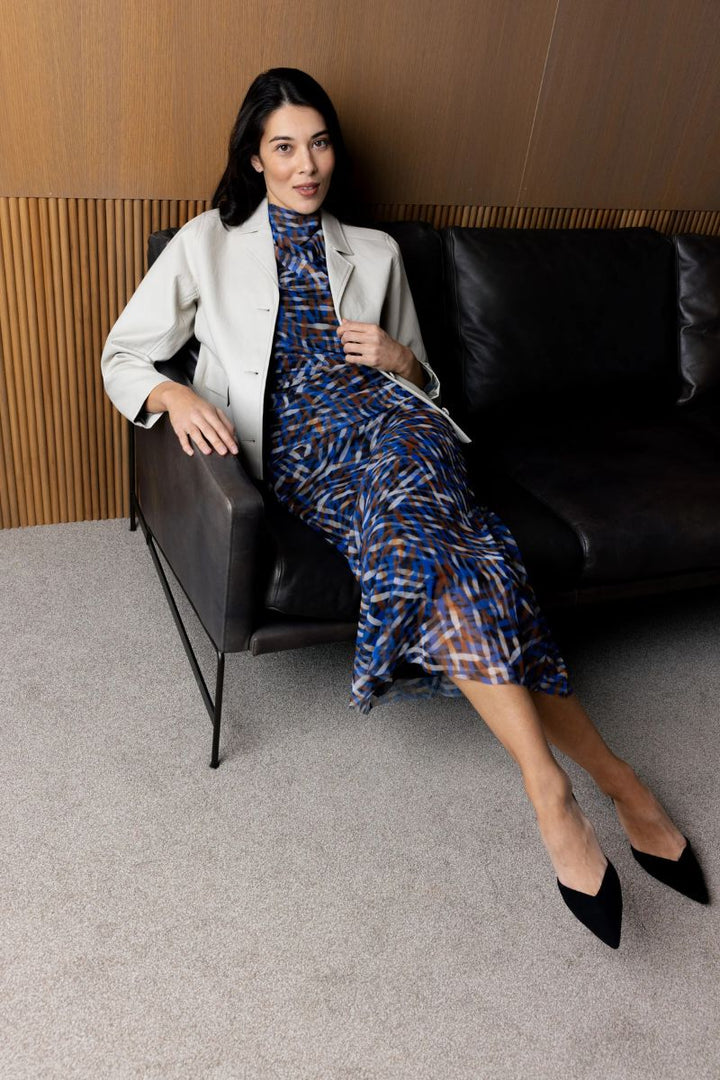 Woman Sitting on a black leather couch wearing a foil clothing blue outfit with the Faux real jacket in Stone.