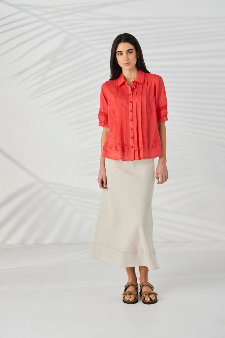 A woman stands in a studio wearing Lania Kristal Shirt in tangelo styled with a  long cream skirt and brown sandals. The background is white with soft leaf shadows.