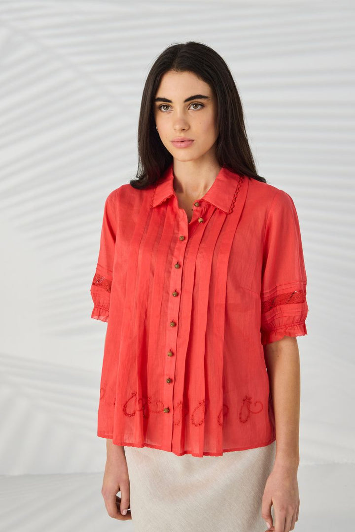 A woman modeling Lania Kristal Shirt in Tangelo paired with a light beige skirt against a white background with subtle leaf shadows.