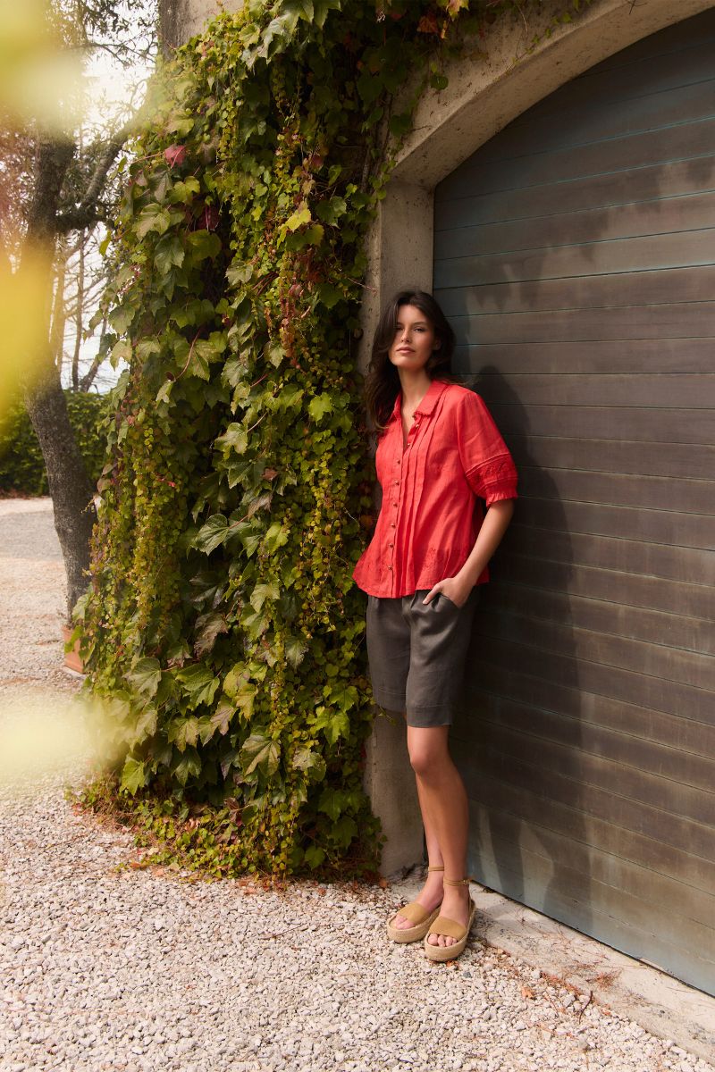 A woman stands outdoors against a gray garage door next to a wall covered in green ivy, wearing Kristal shirt by Lania styled with dark gray shorts and tan sandles.
