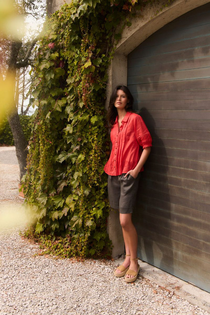 A woman stands outdoors against a gray garage door next to a wall covered in green ivy, wearing Kristal shirt by Lania styled with dark gray shorts and tan sandles.