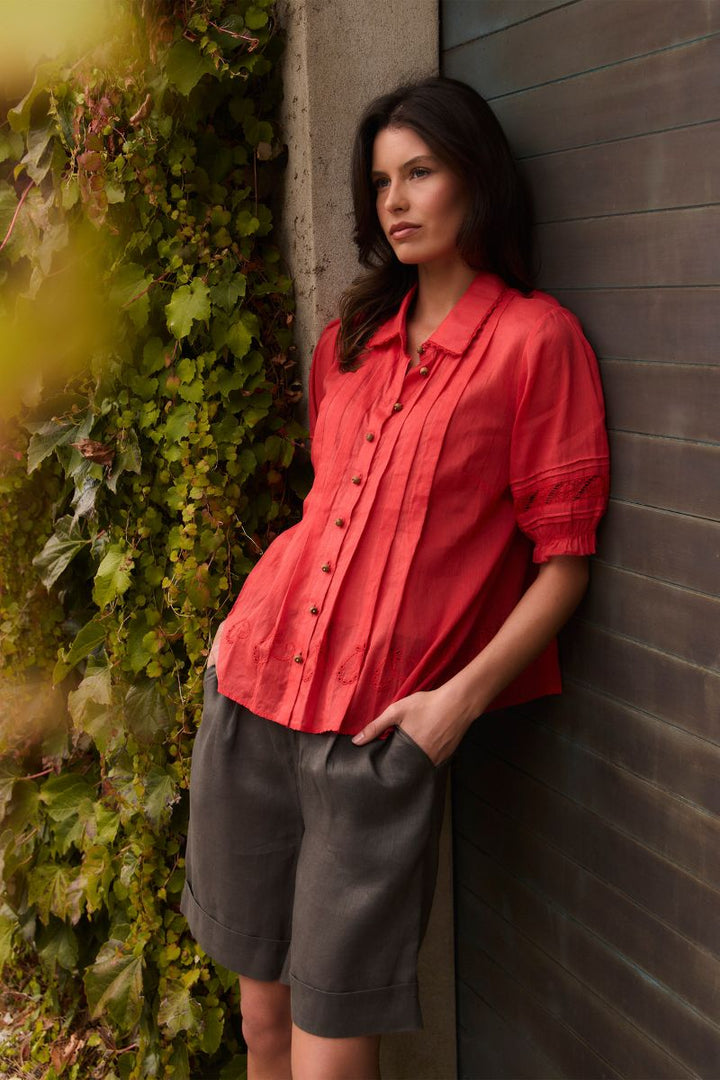 A woman wearing the Lania Kristal Shirt in tangelo styled with dark gray shorts, leaning against a dark wood-paneled wall next to a wall covered in green ivy.