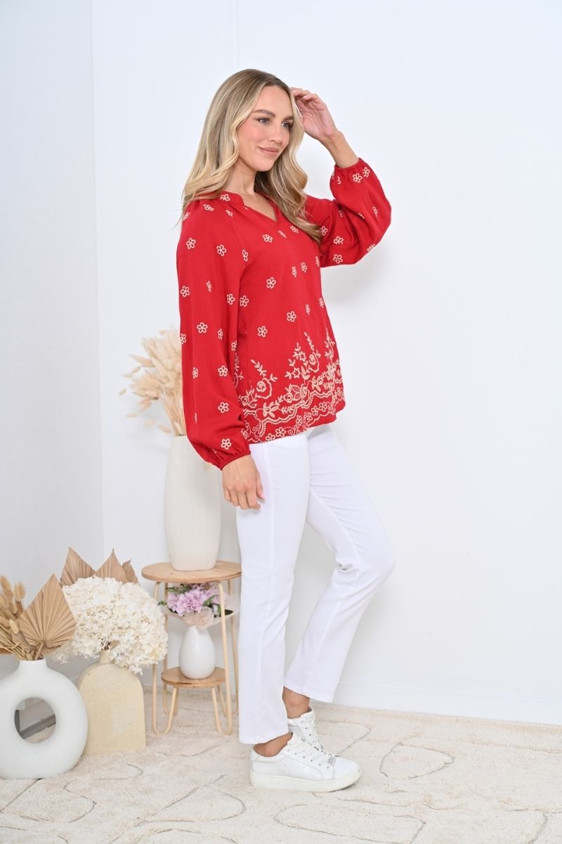 Side view of a Woman wearing a red patterned blouse and white pants standing against a white wall.