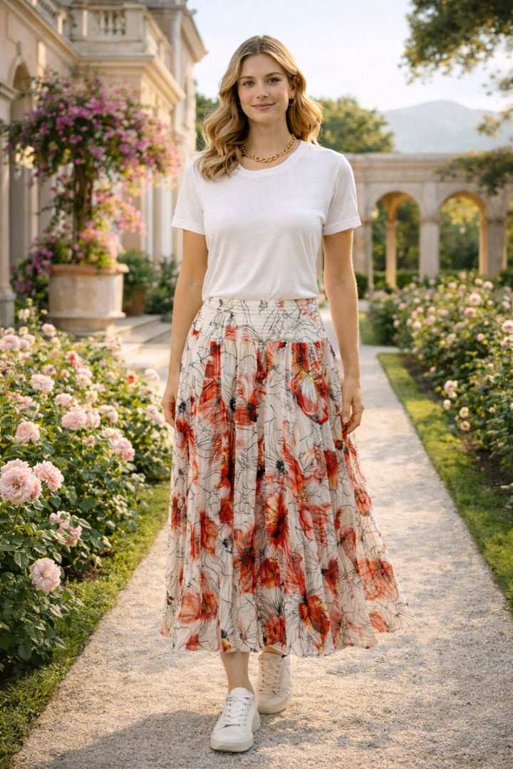Woman in a shirred floral skirt by dress addict with a white top standing in a garden path with flowers and classical architecture.