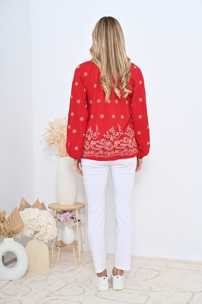 Back view of woman wearing a red embroidered blouse and white pants in a minimalistic setting.